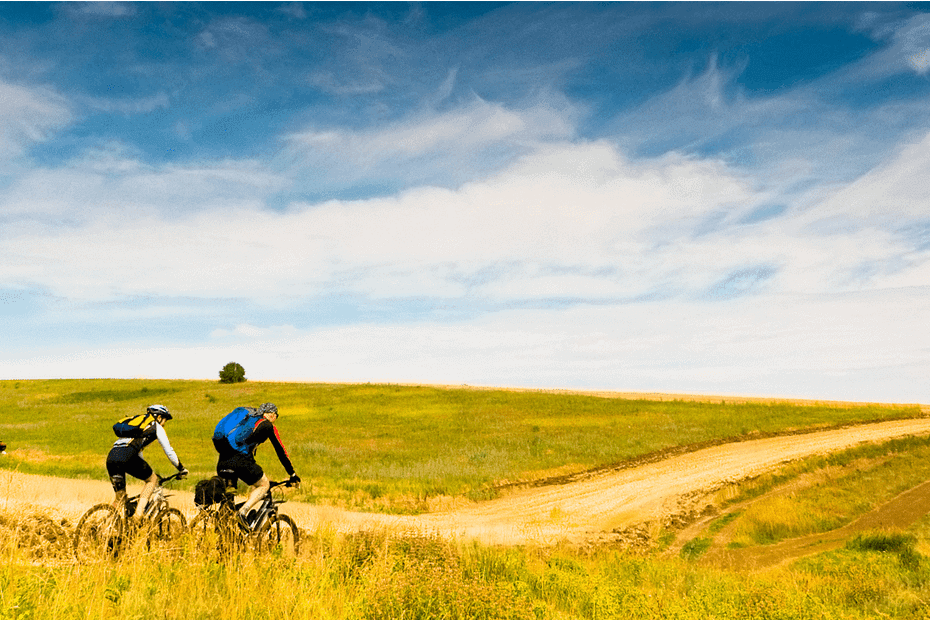 Three people biking on a dirt trail through a field under a cloudy blue sky.
