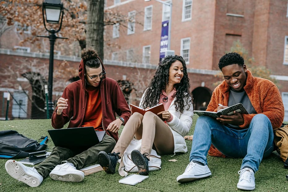 Three employees sit together outdoors on a campus lawn, studying, collaborating, and laughing, illustrating how social connection, learning, and positive workplace relationships contribute to employee well-being.