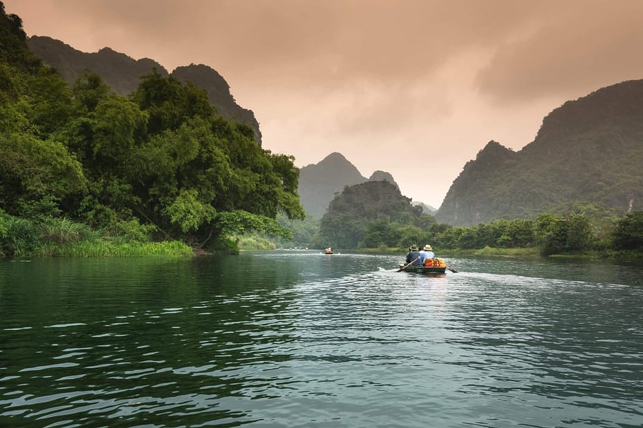 A small boat travels down a calm river surrounded by lush green mountains, representing the strategic journey organizations take to build successful employee wellness programs through steady, long-term health promotion.