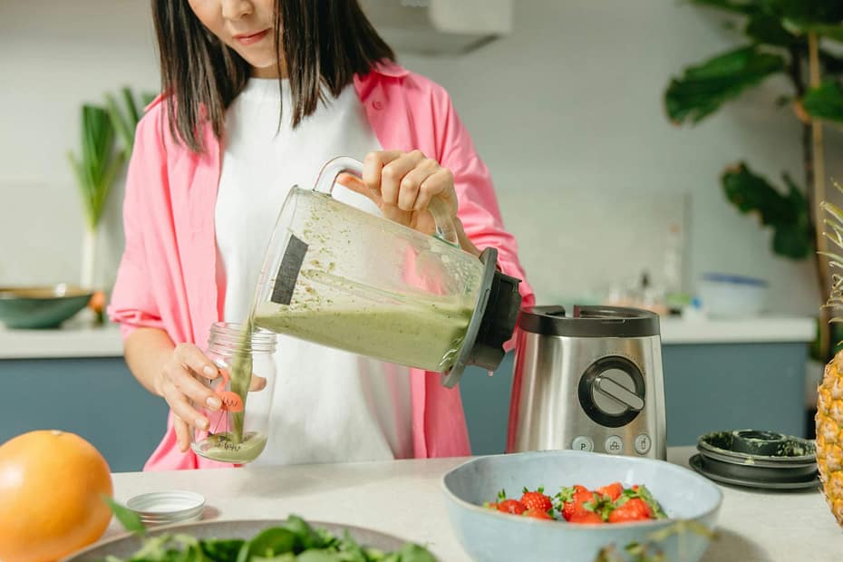 A person pours a green smoothie into a reusable glass bottle in a bright kitchen surrounded by fresh fruits and vegetables, representing healthy nutrition habits supported by workplace wellness programs.