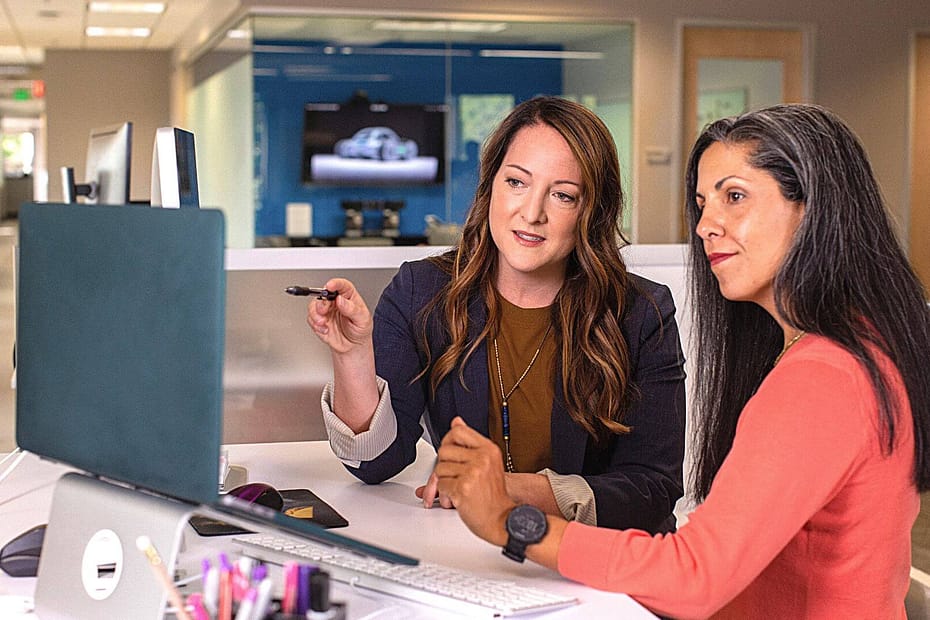 Two women sitting at one desk reviewing something on a blue laptop.