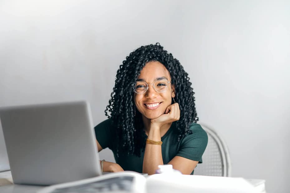 Happy woman sitting at a desk looking into the camera smiling. Employee participating in a digital wellness program on a laptop demonstrates how online wellness platforms provide personalized health coaching, wellness resources, and flexible support for healthier workplace habits.