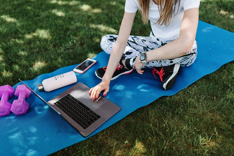 Employee uses a laptop outdoors on a yoga mat with a smartwatch, water bottle, dumbbells, and smartphone, illustrating how digital wellness platforms make workplace health accessible anywhere.