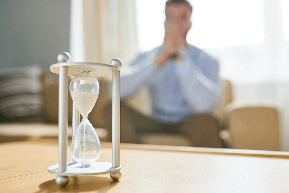 A man sits blurred in a tan office chair while an hourglass in the foreground comes into sharp focus. Employee wellness programs that address stress management and burnout prevention can help employees improve focus, resilience, and overall wellbeing.